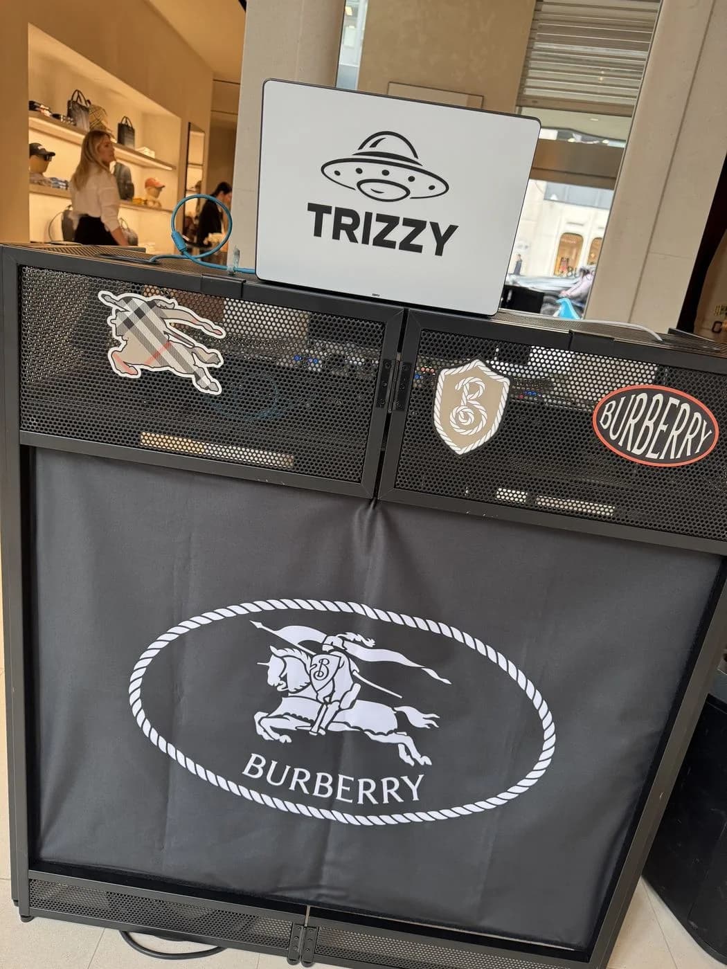 DJ booth wrapped in a Burberry equestrian-logo cover, topped with a TRIZZY sign, inside a Burberry store
